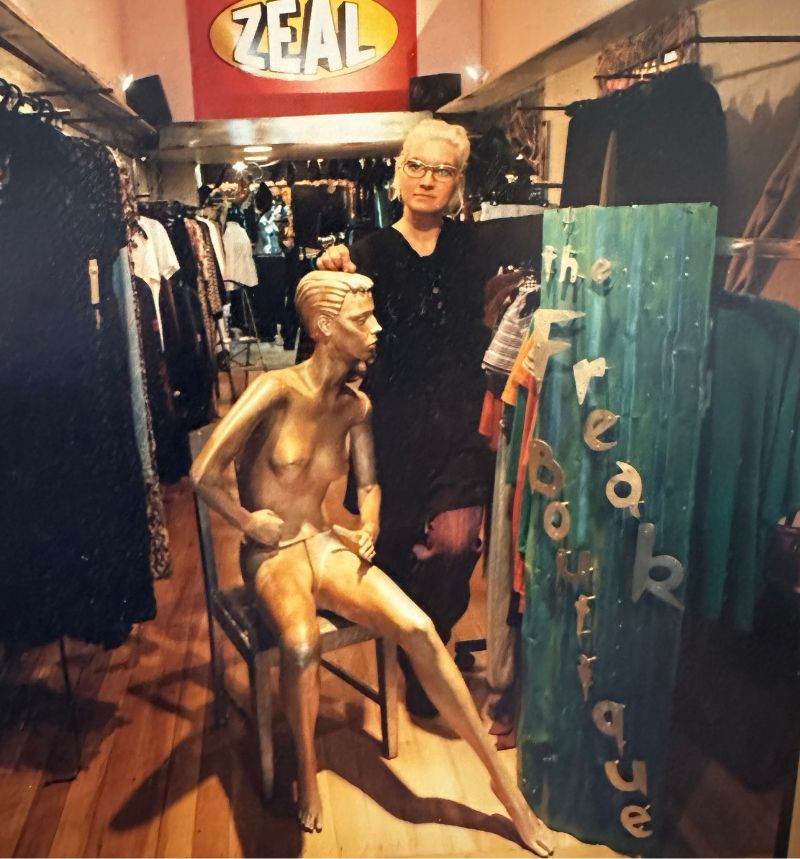 Young woman standing inside a small boutique, surrounded by eclectic clothing racks, a golden mannequin, and a handcrafted metal sign reading “The Freak Boutique.
