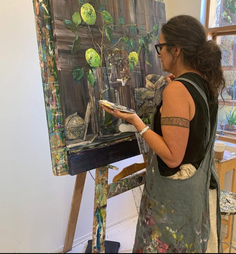 Side view of a woman closely focused on painting green apples and branches in a glass vase, holding a paint palette, with soft light from a nearby window and white walls behind.