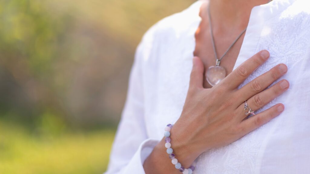 A woman wearing a white blouse places one hand gently over her heart, with only her torso visible.