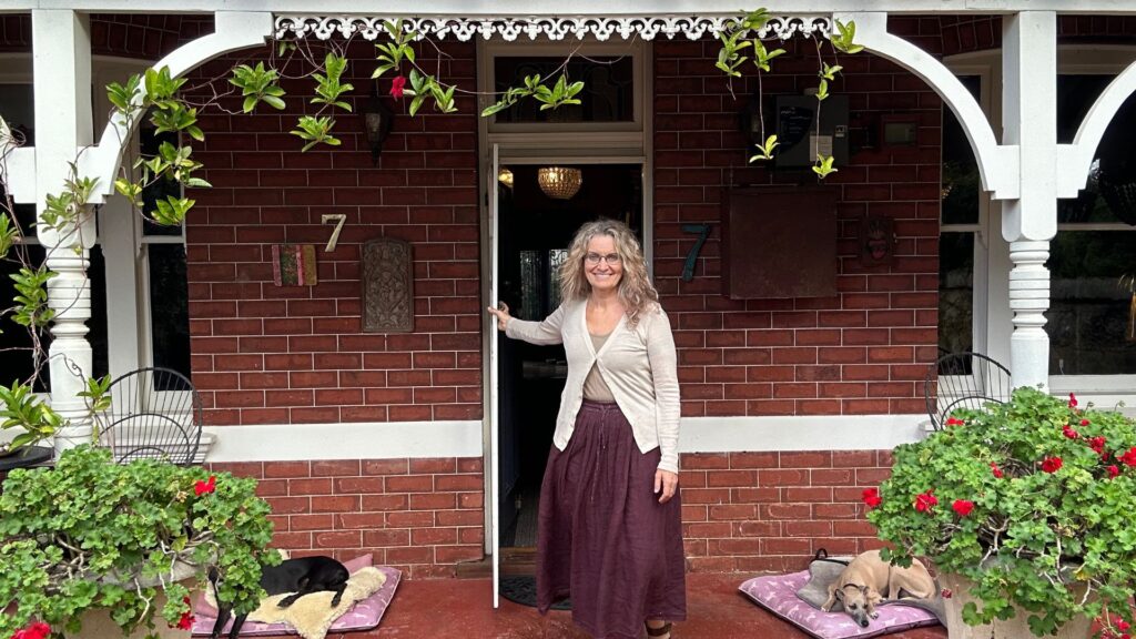 A woman stands barefoot in a sunlit doorway, framed by leafy green plants and two resting dogs on a brick veranda.